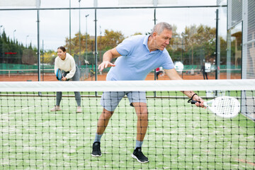 Focused elderly European man pensioner padel player hitting ball with racket on hard court in autumn