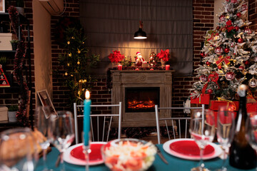 Festive christmas tree near fireplace, home decorated for winter holiday celebration with family at dinner table. Dinning room with fresh food and alcohol glasses for xmas celebration.