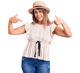 Young beautiful woman wearing summer hat and t-shirt looking confident with smile on face, pointing...