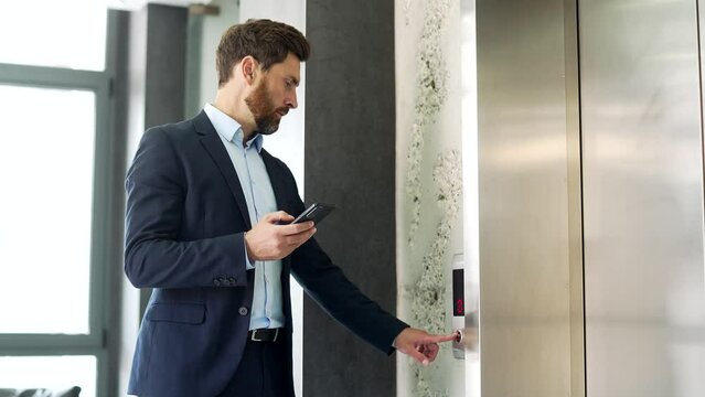 Businessman calling the elevator and browsing using a smartphone in a modern office building. Thoughtful handsome entrepreneur looking at the mobile phone while the lift door opens and goes inside