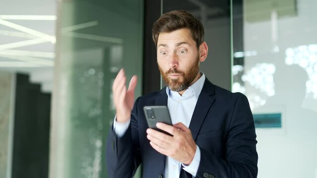 Excited happy businessman in formal suit received great news on smartphone standing at workplace in business office. Joyful entrepreneur reads a pleasant message on a mobile phone, celebrates success