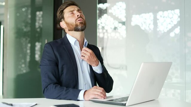 Businessman in a formal suit is hot while sitting at laptop at workplace in business office. The man waves his hands like a fan, cannot concentrate on work, because air conditioner in room is broken