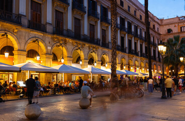 Evening view of bustling Placa Reial in Barcelona with well-lit cafes and people enjoying leisurely...