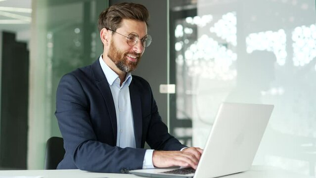 Happy businessman works on a laptop while sitting at workplace in business office. Smiling entrepreneur in formal suit typing massage chatting banking in computer app, using software application