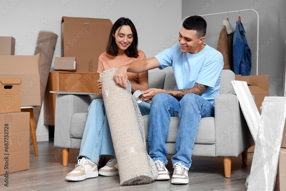 Poster Happy young couple wrapping carpet in room on moving day