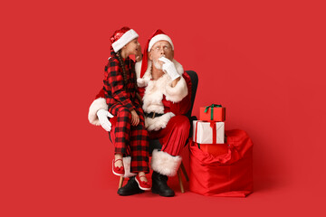 Cute little girl with gift boxes telling Santa Claus her wish on armchair against red background