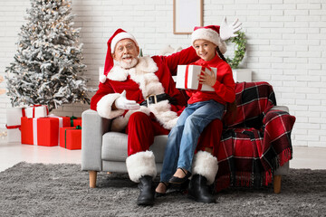 Santa Claus and cute little girl with gift box sitting on sofa in decorated room for Christmas