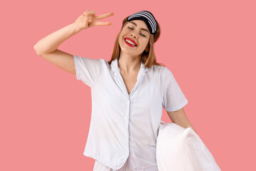 Portrait of pretty young woman in pajamas with sleep mask and pillow on pink background