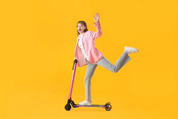 Portrait of excited businesswoman riding kick scooter and waving hand on yellow background - obrazy, fototapety, plakaty