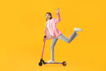 Foto op Plexiglas Portrait of excited businesswoman riding kick scooter and waving hand on yellow background © Pixel-Shot
