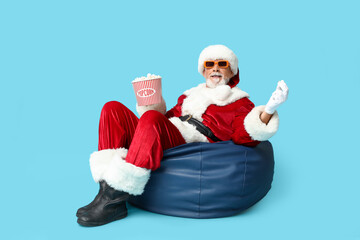 Santa Claus in 3D glasses with bucket of tasty popcorn on blue background