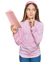 Beautiful brunette little girl reading a book serious face thinking about question with hand on chin, thoughtful about confusing idea