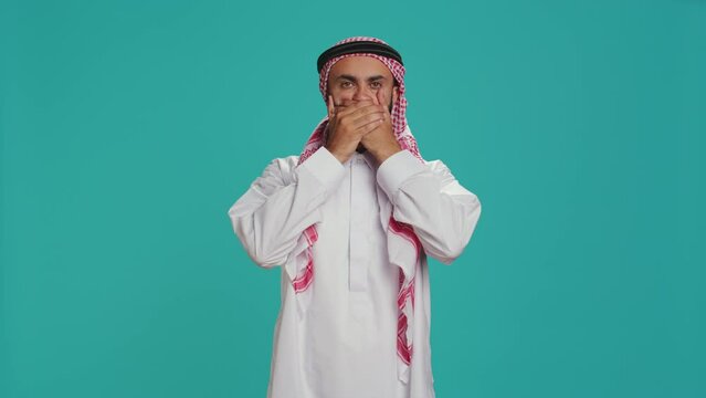 Muslim adult covers eyes, ears and mouth in studio, showcasing three wise monkeys metaphor sign. Middle eastern guy presenting dont hear, see or speak concept symbol in arabic clothes.