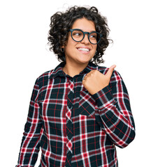 Young hispanic woman with curly hair wearing casual clothes and glasses smiling with happy face looking and pointing to the side with thumb up.