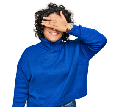 Young hispanic woman with curly hair wearing turtleneck sweater smiling and laughing with hand on face covering eyes for surprise. blind concept. - Powered by Adobe