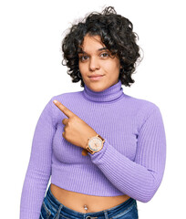 Young hispanic woman with curly hair wearing casual clothes pointing with hand finger to the side showing advertisement, serious and calm face