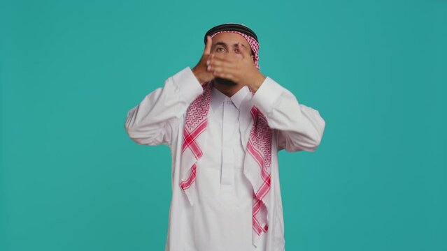 Male model covers eyes, ears and mouth on camera, presenting three wise monkeys symbol. Middle eastern person creating dont hear, see or speak metaphor sign, islamic clothing.