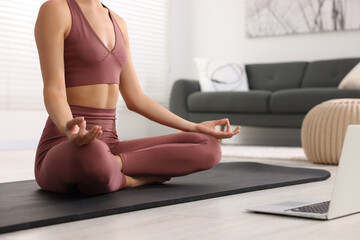 Woman practicing Padmasana with laptop on yoga mat at home, closeup. Lotus pose