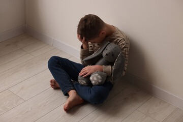 Child abuse. Upset boy with toy bunny sitting on floor near wall indoors