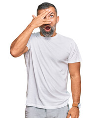 Middle age handsome man wearing casual white tshirt peeking in shock covering face and eyes with hand, looking through fingers with embarrassed expression.