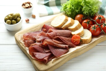 Board with delicious bresaola served with bread and tomato on white wooden table