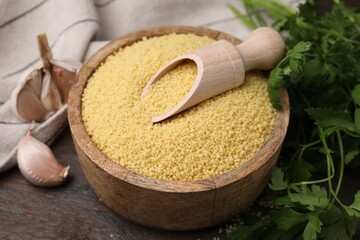 Raw couscous in bowl, scoop, parsley and garlic on table, closeup