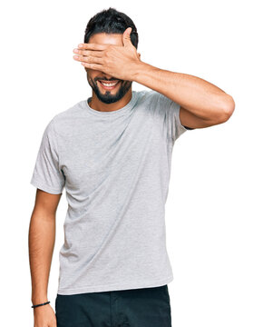 Young man with beard wearing casual grey tshirt smiling and laughing with hand on face covering eyes for surprise. blind concept.
