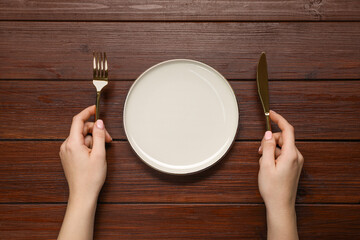 Woman holding fork and knife near empty plate at wooden table, top view - Powered by Adobe