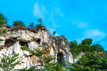 A weathered limestone cliff, adorned with a smattering of trees, stands tall against a clear blue sky in Setigi, Gresik, East Java, Indonesia.