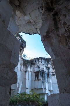 A rectangular opening in a dark, mossy limestone cave frames a bright blue sky with fluffy white clouds in Setigi, Gresik, East Java, Indonesia.