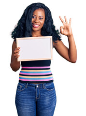 Beautiful african american woman holding blank empty banner doing ok sign with fingers, smiling friendly gesturing excellent symbol