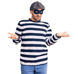 Young handsome man wearing burglar mask clueless and confused with open arms, no idea concept.