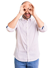 Young handsome man wearing elegant shirt doing ok gesture like binoculars sticking tongue out, eyes looking through fingers. crazy expression.