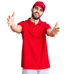 Young handsome man with beard wearing delivery uniform looking at the camera smiling with open arms for hug. cheerful expression embracing happiness.