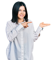 Young brunette woman with blue eyes wearing oversize white shirt amazed and smiling to the camera while presenting with hand and pointing with finger.