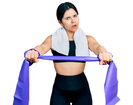 Young brunette woman with blue eyes training arm resistance with elastic arm bands in shock face, looking skeptical and sarcastic, surprised with open mouth