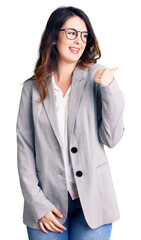 Beautiful young brunette woman wearing business clothes and glasses smiling with happy face looking and pointing to the side with thumb up.