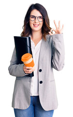 Beautiful young brunette woman holding business folder and drinking coffee doing ok sign with fingers, smiling friendly gesturing excellent symbol