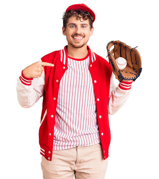 Young handsome man with curly hair wearing baseball uniform holding golve and ball pointing finger to one self smiling happy and proud