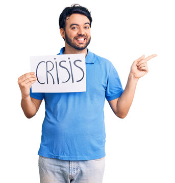 Young hispanic man holding crisis message paper smiling happy pointing with hand and finger to the side