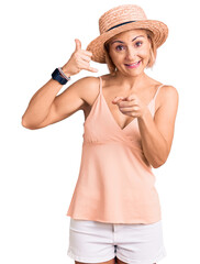 Young blonde woman wearing summer hat smiling doing talking on the telephone gesture and pointing...
