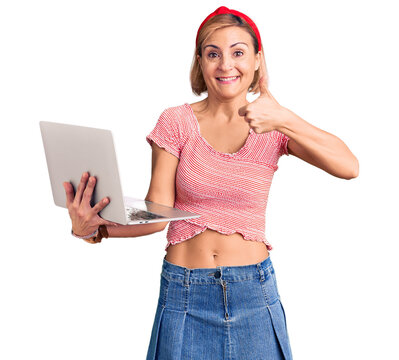Young blonde woman holding laptop smiling happy and positive, thumb up doing excellent and approval sign