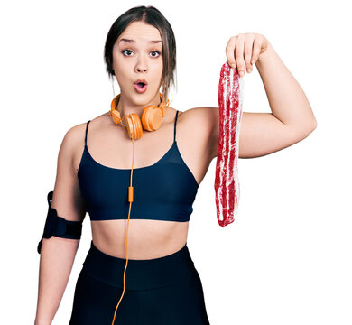 Young hispanic girl wearing sportswear holding meat scared and amazed with open mouth for surprise, disbelief face