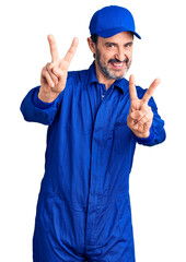 Middle age handsome man wearing mechanic uniform smiling with tongue out showing fingers of both hands doing victory sign. number two.