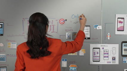 Beautiful asian businesswoman writing Ux and Ui design on board. Project manager looking at board while thinking about project. Woman working by using mind map sharing idea. Back view. Manipulator.