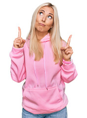 Young blonde woman wearing casual sweatshirt pointing up looking sad and upset, indicating...