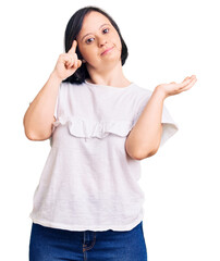 Fototapeta premium Brunette woman with down syndrome wearing casual white tshirt confused and annoyed with open palm showing copy space and pointing finger to forehead. think about it.