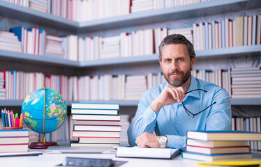 Portrait of teacher with book in library classroom. Handsome teacher in university library. Teachers Day. Teacher giving classes. School teacher in library. Tutor at college library on bookshelf.