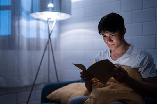 Asian young reader man addict reading a fiction in paperback book while sitting on comfort couch with lamp light after wake up at bedtime unhealthy leisure lifestyle