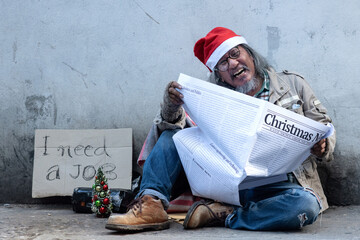 Homeless old man wearing a Santa hat laughs happily when he receives good news in the newspaper,...